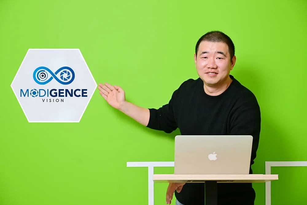 CEO Kim Dae-hee standing at a laptop and gesturing toward the ModigenceVision logo